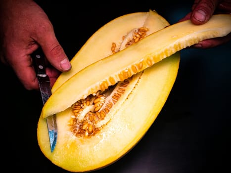 A yellow melon cut with a knife in the hands of a man. Food photo. Melon culture. Agriculture. Place for your text. Black background. Background image. Shchablon. Poster. Vegetarian food.