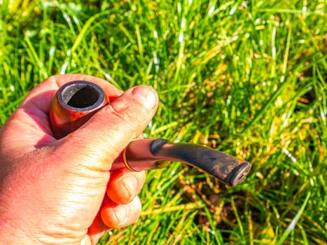 Wooden smoking pipe for tobacco in hand. Pipe for smoking tobacco. Smoker's tool. Human hand. Bad habits. Nicotine addiction. Nicotine in cigarettes. Green grass. Background image. Poster.