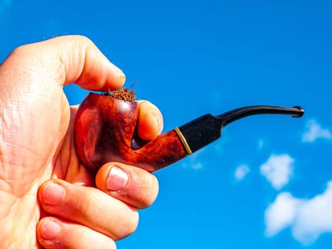 Wooden smoking pipe for tobacco in hand against blue sky background. Pipe for smoking tobacco. Smoker's tool. Human hand. Bad habits. Nicotine addiction. Nicotine in cigarettes. Background.