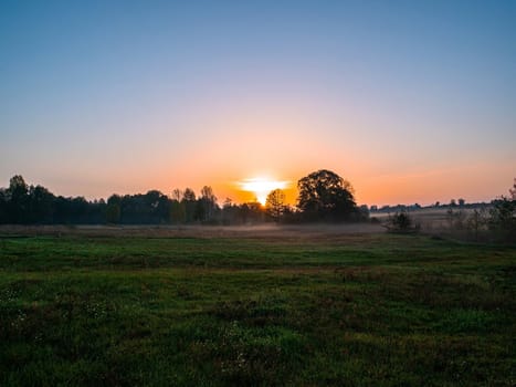 Orange sunrise over a meadow in fog. Orange sunny sunset over the horizon. Trees on the horizon. Morning fog over the meadow. The morning dawn of the rising sun. Evening dawn of the sunset.