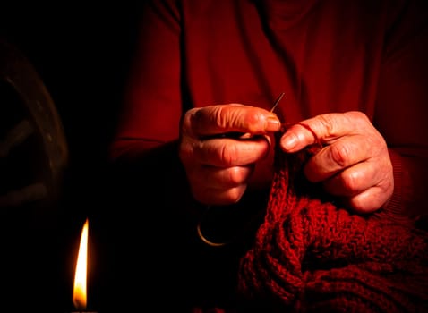 Hand knitting with woolen thread by candlelight. Knitting needles. Woolen thread. Old man's hands. Handwork. Weave warm clothes. Manual creativity. Self made. The flame of a wax candle.