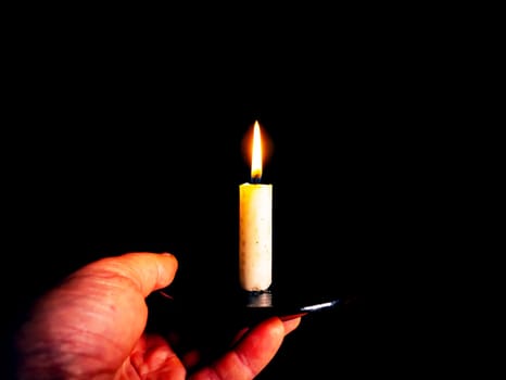 The flame of a burning wax candle in the hands of a man. The flame of a wax candle. Fire in the dark. Day of Remembrance. A minute of silence. Funeral services. Sorrows on Memorial Day. Night.