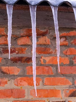 Icicles of frozen water on the roof of a house with a brick wall. Snow melting. Winter. Spring drops. Ice icicle. Frozen water. Brick wall. Frosty frost. Winter season. Roof of the house.