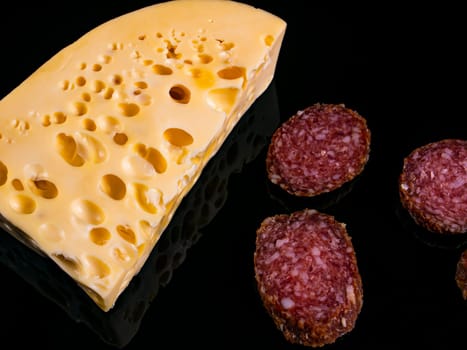 A piece of hard cheese and a sliced sausage on a black background. Hard cheeses. Meat sausage. Milk product. Meat sausages. Dairy products. Home kitchen. Food photo. Black background.