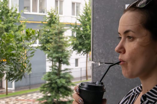 One black paper cup with a lid and a straw in the hands of a young happy smiling woman outdoors in summer in sunny weather on the background of houses, in a coffee shop or restaurant. Coffee to go