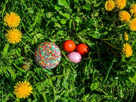 Colored Easter eggs in green grass with dandelion flowers. Easter eggs. Blooming yellow dandelions. Green grass. Christian holiday. Religious tradition. Christian culture. Spring season.