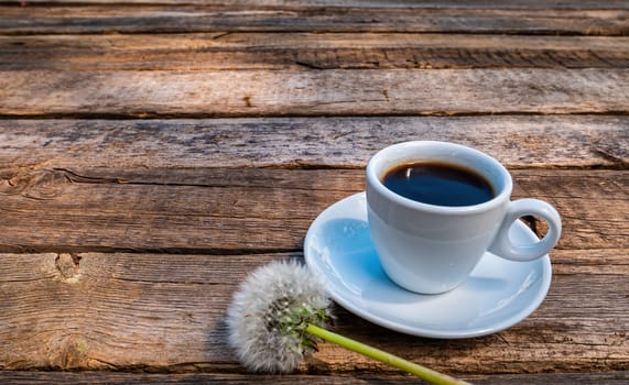 Cup of coffee and dandelion flower on a wooden table. Coffee drink. Dandelion flower. Cup and saucer. Wooden table from a bar. Food and drinks. Ceramic tableware. Street Cafe.