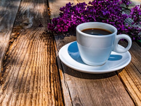 A cup of coffee and a blossoming branch of lilacs on a wooden table. Coffee drink. Lilac tree flowers. Cup and saucer. Wooden table from a bar. Food and drinks. Ceramic tableware. Street Cafe.