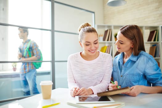 Friendly teenage girls sitting by desk while studying on background of groupmate