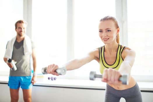Young female exercising with dumbbells with guy on background