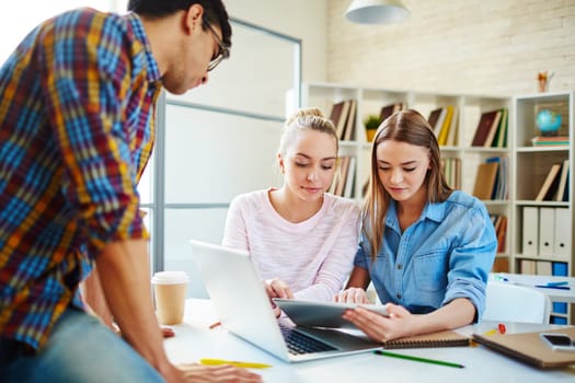 Two cute teenage girls looking through data in touchpad with their groupmate near by