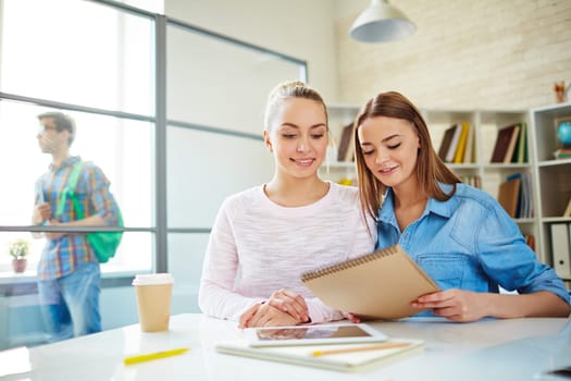 Friendly teenage girls reading notes by desk with their groupmate on background