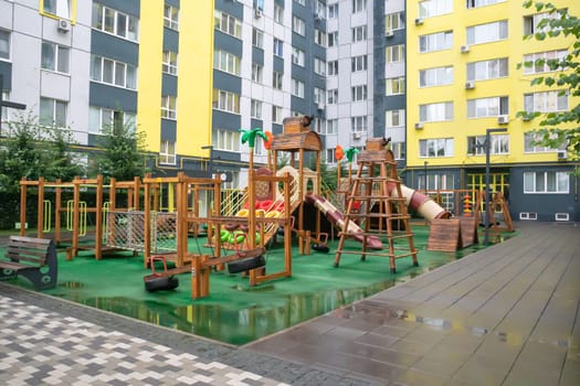 A courtyard of high-rise buildings with a modern and large playground made of wood and plastic on a rainy summer day without people. Empty outdoor playground. A place for children's games and sports