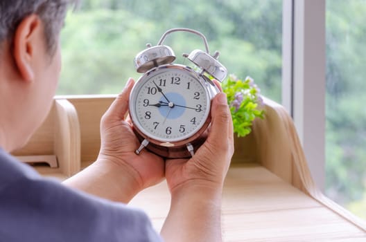 Senior man sitting at desk and holding white alarm clock