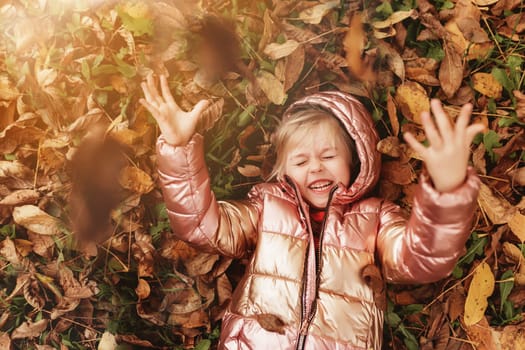 Happy little blonde girl plays with yellow autumn leaves in the garden. The child smiles and has fun in park. Playful kid throwing up leaves in autumn season park. Child lays on the ground.