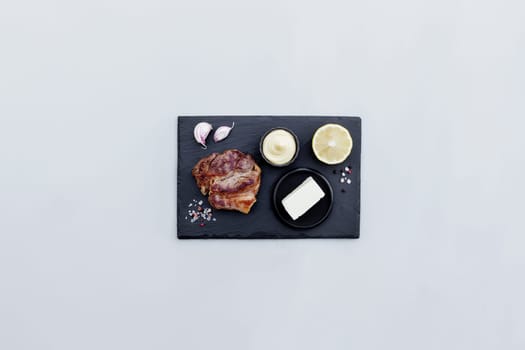 Gray concrete background flat lay with cut of medium-rare red ribeye meat steak on black cutting board, lemon, pepper seasoning, salt, mayonnaise sauce, garlic, butter. Rib-eye fillet with copy space