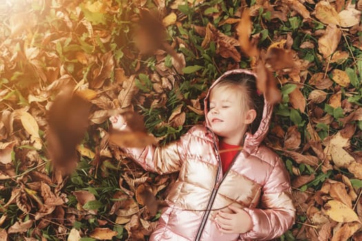 Happy little blonde girl plays with yellow autumn leaves in the garden. The child smiles and has fun in park. Playful kid throwing up leaves in autumn season park. Child lays on the ground.