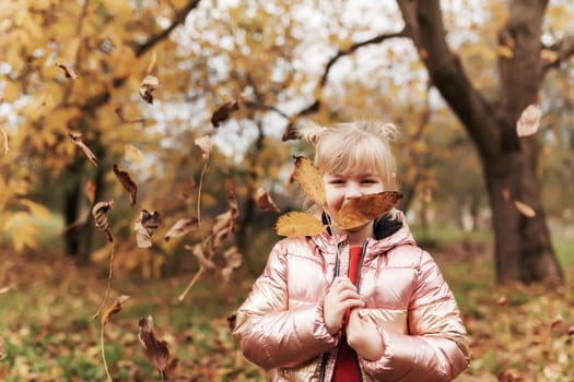 Happy little blonde girl plays with yellow autumn leaves in the garden. The child smiles and has fun in park. Playful kid throwing up leaves in autumn season park. Child lays on the ground.