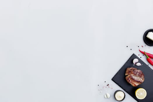 Gray concrete background flat lay with cut of medium-rare red ribeye meat steak on black cutting board, lemon, pepper seasoning, salt, mayonnaise sauce, garlic, butter. Rib-eye fillet with copy space