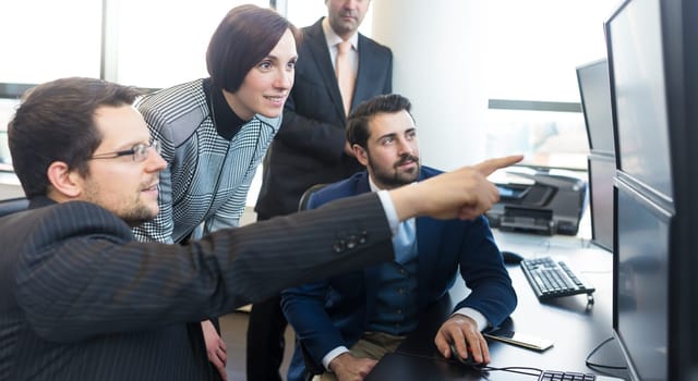 Business team looking at data on multiple computer screens in corporate office. Businessman pointing on screen. Business people trading online. Business, entrepreneurship and team work concept.