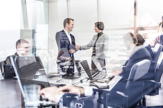 Sealing a deal. Business people shaking hands, finishing up meeting in corporate office. Businessmen working on laptop seen in glass reflection. Business and entrepreneurship concept.
