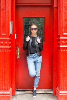 Fashionable Young Woman Wearing Sunglasses, Black Leather Jacket and Bleu Jeans Posing in front of Red Door on Street of New York City
