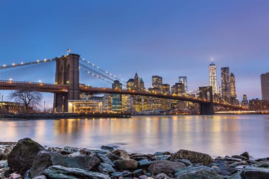Brooklyn bridge and New York City Manhattan downtown skyline at dusk with skyscrapers illuminated over East River panorama.