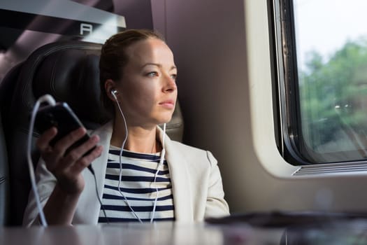 Thoughtful businesswoman looking trough the window, listening to podcast on cellphone using headphone set while traveling by train in business class seat.