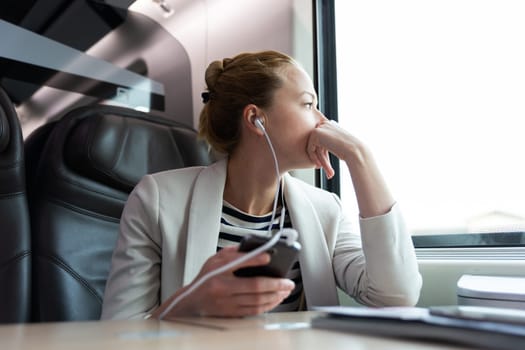 Thoughtful businesswoman looking trough the window, listening to podcast on cellphone using headphone set while traveling by train in business class seat.