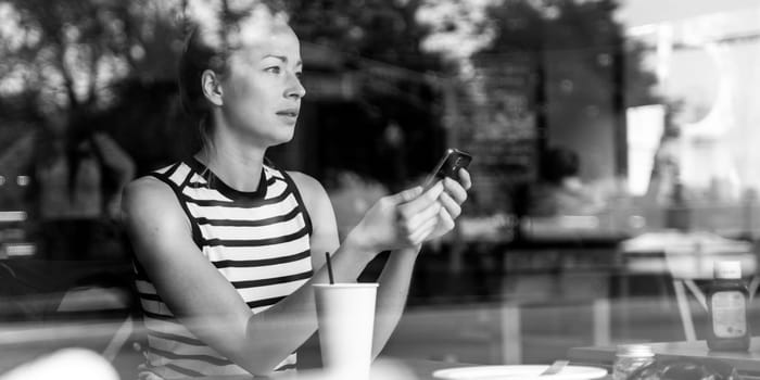 Thoughtful caucasian woman holding mobile phone while looking through the coffee shop window during coffee break. Street reflections in the window glass. Black and white image.