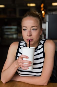 Young caucasian woman sitting alone in coffee shop drinking american coffee, people watching, thoughtfully looking trough the coffee shop window.