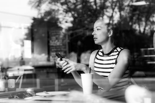 Thoughtful caucasian woman holding mobile phone while looking through the coffee shop window during coffee break. Street reflections in the window glass. Black and white image.