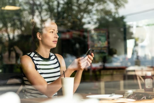 Thoughtful casual caucasian woman holding mobile phone while looking through the coffee shop window during coffee break. Street reflections in the window glass.