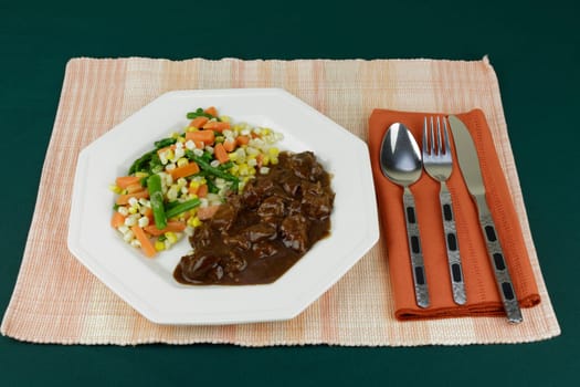Mixed steamed vegetables meal of green asparagus, orange carrots and yellow and white corn with beef tips in gravy. Gravy covered beef tips with and mixed vegetables lunch on a white octagon plate.