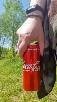 Hand holding a can of Coca Cola. The drinks are produced by the American beverage corporation The Coca-Cola Company. Young woman with Coca-Cola outdoors, close-up. Ukraine, Kiev - August 19, 2021