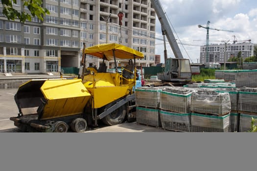 An asphalt paver is parked at a construction site. Road construction machinery stands by the road in a warehouse. Construction of heavy roads and repair of city streets