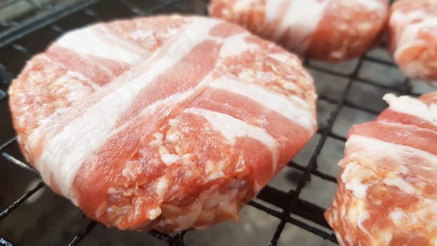 Raw meat cutlets wrapped in bacon are cooked on the grill rack. The chef prepares the grilled burger patties. Ingredients for making a hamburger. Meat lovers concept. Fast food