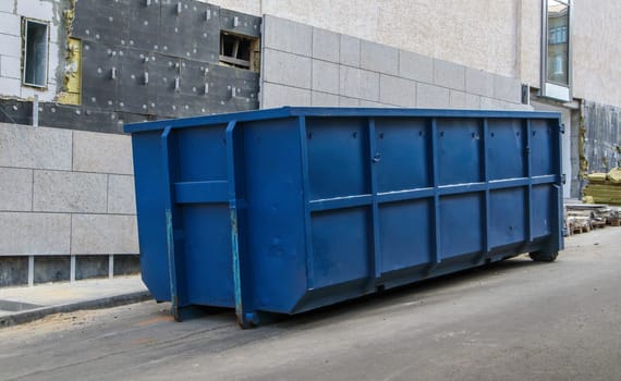 Metal durable blue industrial trash bin for outdoor trash at construction site. Large waste basket for household or industrial waste. A pile of waste