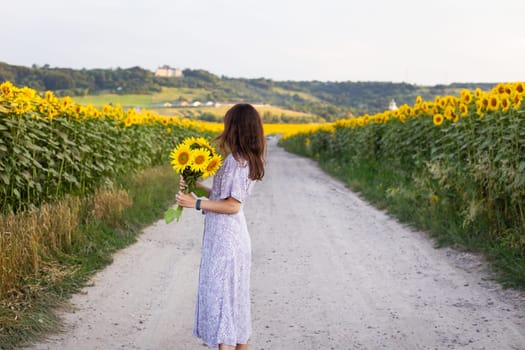 A beautiful young girl in a dress holds a bouquet of sunflowers in her hands stands on the road among a large field with sunflowers