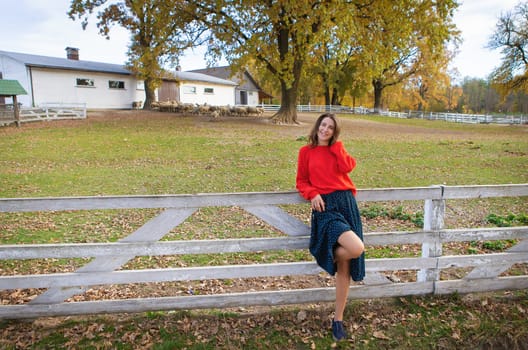 A beautiful girl in a red sweater stands near a white wooden fence. Beautiful warm autumn, interesting weekend, outdoor recreation.