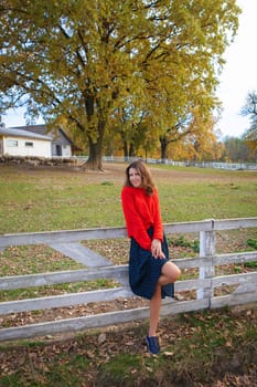 A beautiful girl in a red sweater stands near a white wooden fence. Beautiful warm autumn, interesting weekend, outdoor recreation.