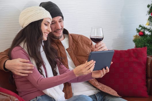 Smiling young couple resting and watching video on sofa in living room. Loving husband embracing wife and holding a glass of red wine while woman holding digital tablet PC. Enjoy weekend time together concept.