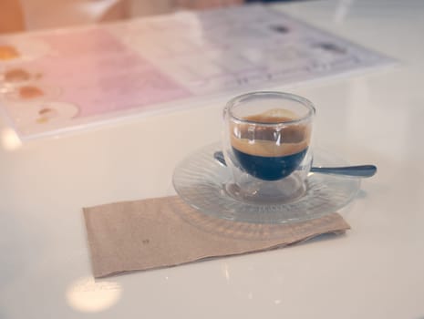 Cappuccino served in a transparent double wall glass cup, on a saucer with a spoon and brown napkin. Hot coffee on white table.