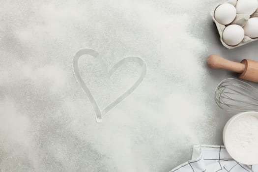 Heart shape from flour on grey concrete background flat lay, copy space. Baking ingredients, kitchen utensils, cooking hobby concept. Prepare dough, egg, bowl flour, rolling pin, whisk, kitchen towel
