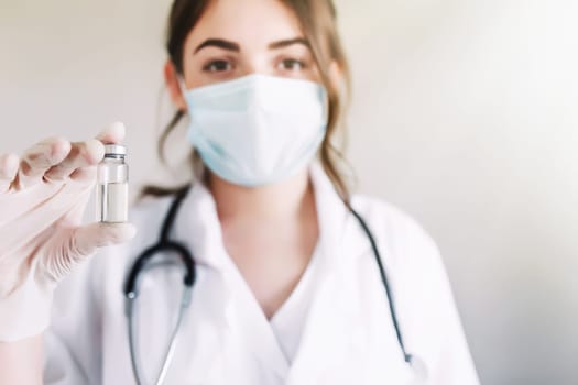 Woman doctor or nurse in uniform and white gloves wearing face mask protective in lab, holding medicine liquid vaccine vial bottle preparing for injection with COVID-19 Coronovirus vaccine for patient