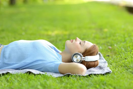 Side view portrait of a relaxed woman listening to music with headphones lying on the grass in a park