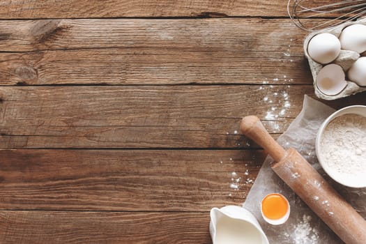 Baking ingredients, kitchen utensils on old wooden background. Cooking dough, preparing egg yolk, flour, rolling pin, milk, parchment paper, whisk, salt, sodium. Concept flat lay photo with copy space