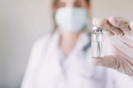 Woman doctor or nurse in uniform and white gloves wearing face mask protective in lab, holding medicine liquid vaccine vial bottle preparing for injection with COVID-19 Coronovirus vaccine for patient