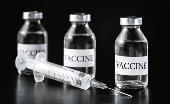 Three glass vaccine vials on black board, hypodermic syringe near.