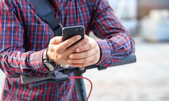 Young man wearing shirt leaning on electric scooter handlebar, holding mobile smartphone in his hands, closeup detail.